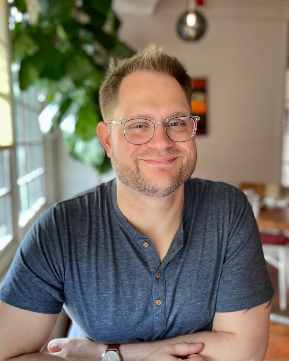 Matthew Hanson, smiling, wearing glasses and a blue shirt, seated at a table by a sunlit window